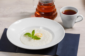 Rice milk porridge in a white plate with butter and mint leaves on a wooden background with a mug of tea. Healthy Breakfast, proper morning.