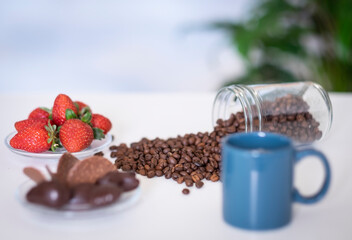 Defocused blue coffee cup and coffee beans on the white table - red strawberries and some chocolate on background