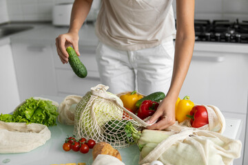 Woman came back from a market and unpacks a reusable grocery bag full of vegetables on a kitchen at home. Zero waste and plastic free concept. Girl is holding mesh cotton shopper with vegetables.