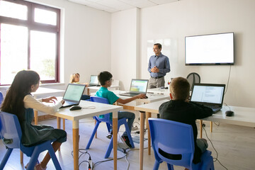 Confident teacher explaining lesson to pupils. Multiethnic children sitting at table in classroom, listening middle-aged man and using laptop computers. Childhood and digital education concept