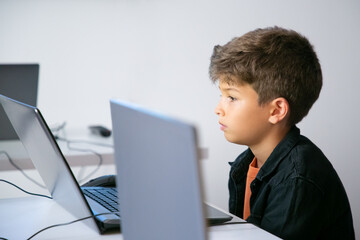 Serious pupil sitting at table and looking at laptop screen. Concentrated school boyreading task or...