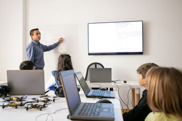 Middle-aged teacher explaining lesson to pupils and writing on board. Children sitting at table with laptop computers in classroom and listening tutor. Childhood and digital education concept