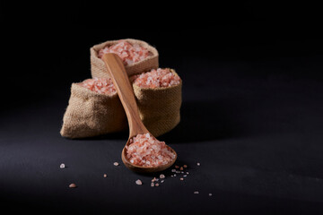 Himalayan salt, thick and pink, in three jute bags along with a wooden spoon with salt. All on a black background.