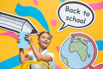 Schoolgirl holding book near speech bubble with back to school lettering and paper art on yellow background