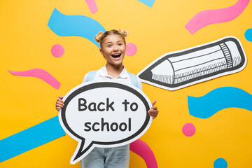 excited schoolchild holding speech bubble with back to school lettering near multicolored paper elements and pencil on yellow