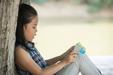  little asian girl reading a book under big tree. children and science. blurred background. learning the imagination and dreams of rural child. studying at home.