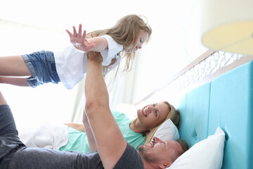 Fototapeta premium Man and woman lie side by side in bed and look up at their daughter. Dad hold girl outstretched arms up. Child rest directly on man arms with his arms out to sides.