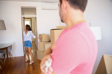 Young man holding key from his new apartment, standing with arms folded and watching his girlfriend carrying carton boxes and looking around. Cropped shot, closeup. Property buying or mortgage concept