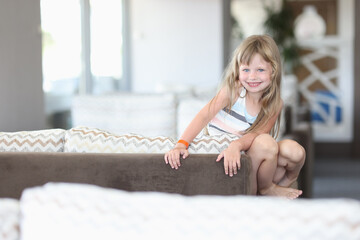 Naklejka premium Beautiful girl in striped dress is squatting on sofa. Child play in room.