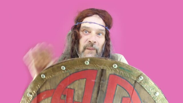 Joke humor. Mature man is surprised and happy, crazy Viking with long hair and beard greets with his hand hiding behind shield. Pink background