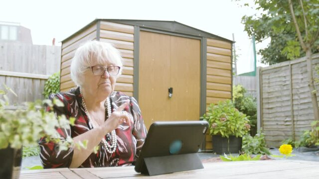 Elderly Woman speaking to a tablet device using her hands and pointing in her garden wide shot
