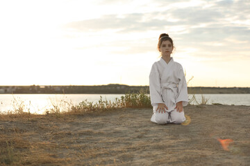 Cute little girl in kimono meditating outdoors at sunset. Karate practicing