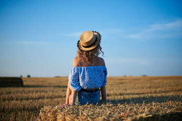Young curly blond woman, wearing jeans shorts, light blue shirt and straw hat, sitting with back to...