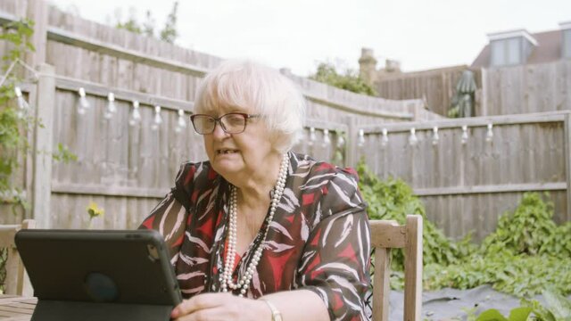 Elderly Woman using Skype on a tablet device tapping on screen while talking, laughing and checking watch in garden