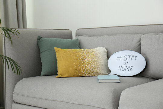 Book and speech bubble with hashtag STAY AT HOME on sofa indoors. Message to promote self-isolation during COVID‑19 pandemic