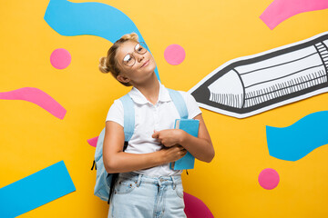dreamy schoolgirl in eyeglasses looking away while holding book on yellow background with paper pencil and abstract elements
