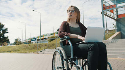 Young caucasian woman in the wheelchair using laptop. Train passing in the background. High quality...