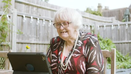 Elderly Woman using Skype waves to tablet device, laughs and speaks in garden in the day