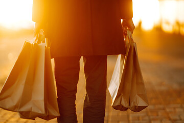 Shopping bags in the hands at sunset. Hand of young man with paper bags with purchases. Consumerism, purchases, shopping, black friday, lifestyle concept.