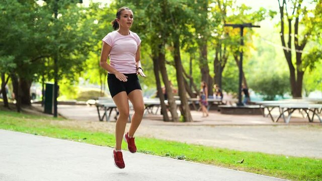 Sporty woman running in place and warming up feet before jogging in park, blurred people on background. Young fit female doing sports outside. Concept of fitness