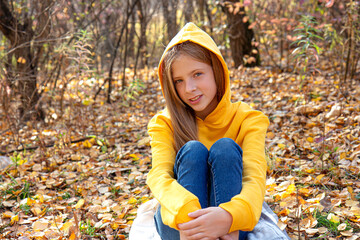 Beautiful teen blonde girl in a bright orange sweatshirt and jeans smiling in an autumn park. Autumn portrait in the forest against the background of fallen golden leaves