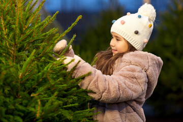 winter holidays and people concept - happy smiling little girl choosing christmas tree at street market in evening