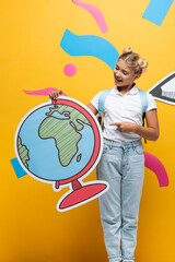 Fototapeta premium excited schoolgirl pointing with finger at globe maquette near paper cut pencil and colorful elements on yellow