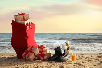 Santa's boots, cocktail, sunglasses and bag with presents on beach. Christmas vacation
