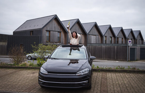 Happy Woman Standing Out Of Car Sunroof. Road Trip Joy. Beautiful Hipster Caucasian Girl Holding A Bottle Of Wine In Her Hands. Carefree Lifestyle