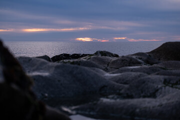 Compressed perspective shot over rocks on the beach shore at dusk looking out towards the horizon. Dark rocks in foreground.
