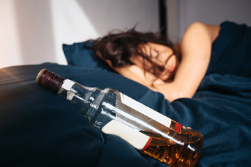 Young beautiful woman in morning bed at home. Sleeping after alcohol party. Bottle of whiskey on bed besides. Sun shines in room.