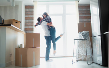Young couple first time home owners celebrate moving day concept, man husband lifting holding wife standing near boxes in new own house apartment, relocation and family mortgage