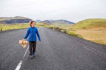 Obraz premium Cute child, preschool boy with teddy bear in hand, running on a road in Iceland on a rainy day