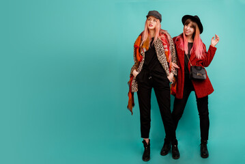 Fototapeta premium Full height image of two happy cheeky girls , best friends posing on blue background. Wearing stylish casual fur coat with leopard print and hat. Space for text.