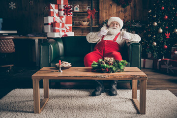 Full length photo jolly grey beard santa claus sit comfort couch enjoy watch x-mas christmas movie fairytale drink milk eat cookies wear red cap headwear in noel advent orament house indoors