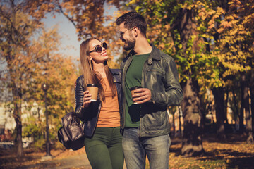 Positive romantic couple students walk in fall october town park hold takeout coffee mug beverage wear sunglass jacket