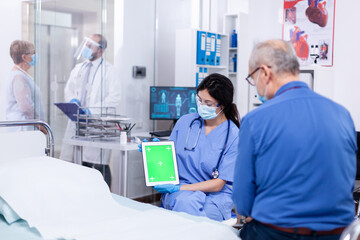 Patient looking at tablet with green screen during consultation in hospital room with nurse wearing face mask against coronavirus as safety precaution. Medical examination for infections, disease .