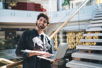 Cheerful Turkish software developer with modern netbook in hands standing indoors and laughing, happy Middle Eastern small owner waiting for delivery newsletter using laptop computer for browsing