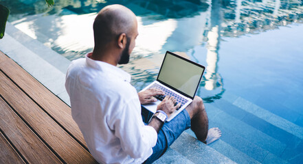 Skilled digital nomad user sitting at pool terrace in courtyard of own Indonesian villa and typing...