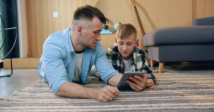 Attractive confident modern dad teaching his teen son how to use i-pad while they spending their free time at home,lying on the carpet