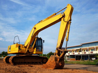 Yellow tracked excavator at construction site. Parked on a ground floor with townhouse background and blue sky with white clouds, heavy equipment concept. Selective focus