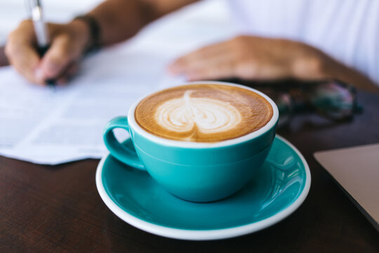 Cropped image of man writing on paper filling in application at working place with aroma hot caffeine beverage, selective focus on coffee mug with cappuccino standing on blurred background