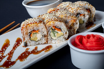Sushi Sets Uramaki, California, Philadelphia, on a white plate. Nearby ginger and wasabi. Soy sauce in a white bowl. On a dark background.
