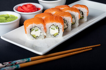 Sushi Sets Uramaki, California, Philadelphia, on a white plate. Nearby ginger and wasabi. Soy sauce in a white bowl. On a dark background.