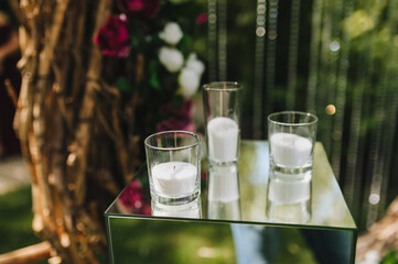 A glass candlestick with a candle, white sand, paraffin and wax inside stands on a glass table. Wedding and party decorations at the ceremony.