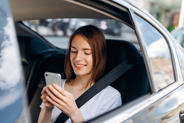 Fototapeta na wymiar Beautiful, young woman uses a smartphone and smiles while sitting in the back seat of a car