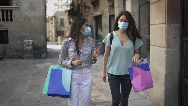 Two young women friends having a walk in the city while shopping with sanitary masks during the coronavirus pandemic. Covid-19 lifestyle, girls with bags having fun.