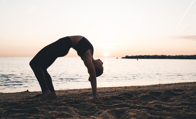 Flexible relaxed woman standing in Wheel pose during evening yoga workout at beach seashore, inspired motivated fit girl in sportswear training body during stretching exercising at coastline