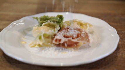 Tortellini on white plate on wooden table, pouring parmesan. Close up tortellini of different colors, green, white, red served with spoon on white plate with melting butter