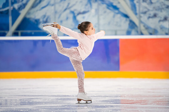 little girl figure skater in light pink tracksuit with smile skates on the ice on an indoor skating rink.